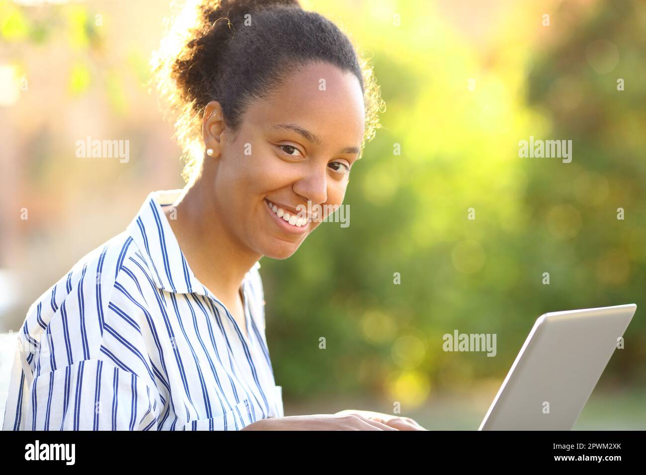 Happy black woman using laptop looking at you sitting in a park Stock Photo