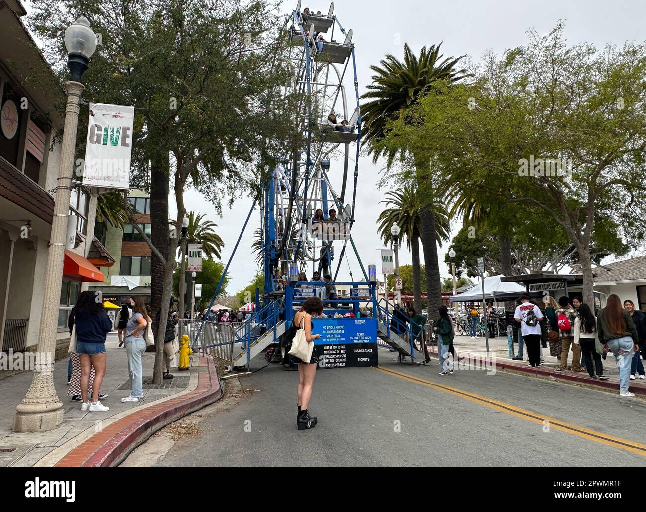 Isla Vista, California, USA. 30th Apr, 2023. Pardall Carnival In Isla Vista (Goleta) California brings together UCSB students, families and community members to promote University of California Santa Barbara student organizations and wellness. A sign Says 'Give'' and a woman in shorts stands in front of a Ferris wheel. (Credit Image: © Amy Katz/ZUMA Press Wire) EDITORIAL USAGE ONLY! Not for Commercial USAGE! Credit: ZUMA Press, Inc./Alamy Live News Stock Photo