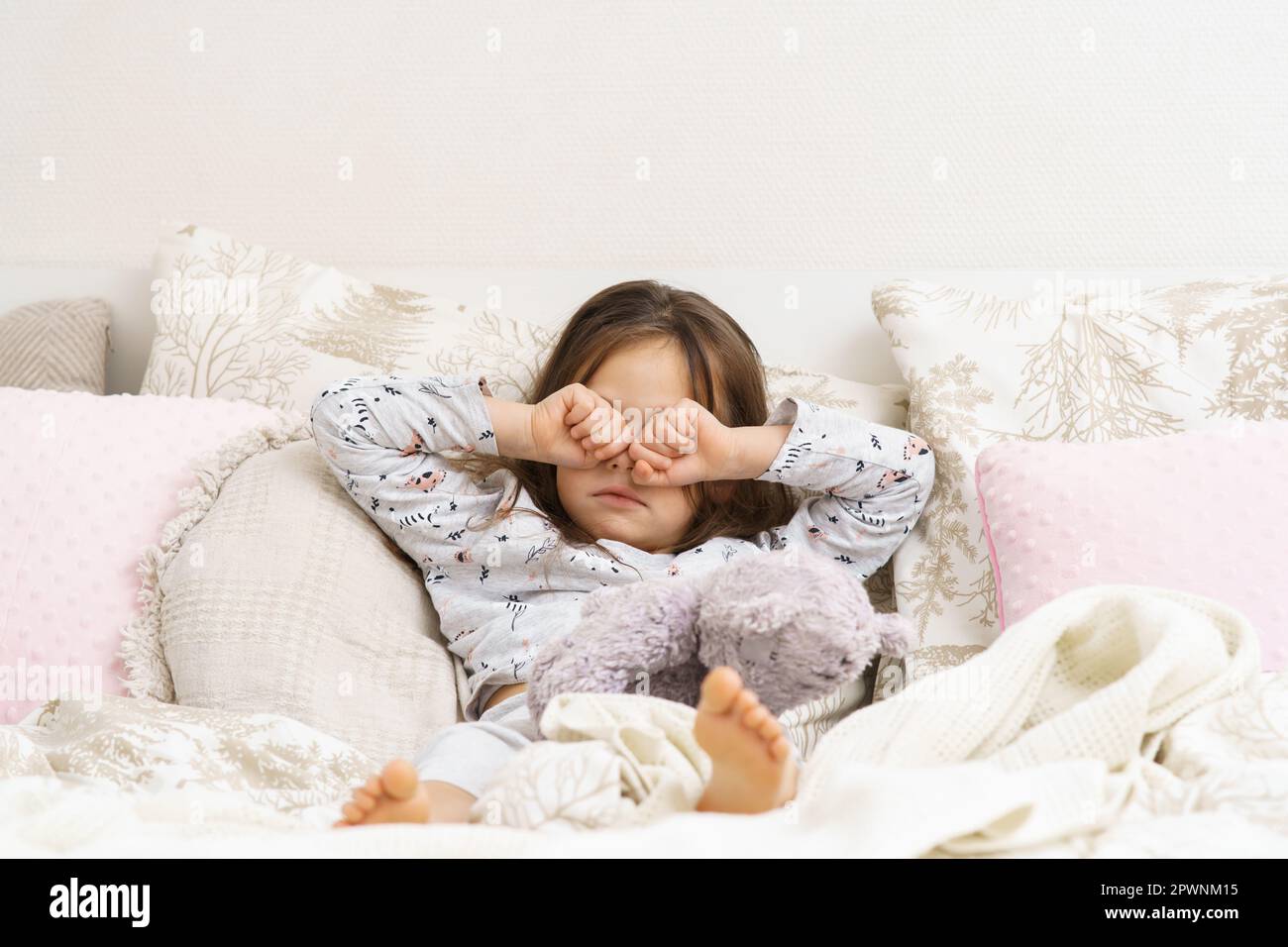 Adorable little girl in sleepwear lying on bed and rub eyes with fists