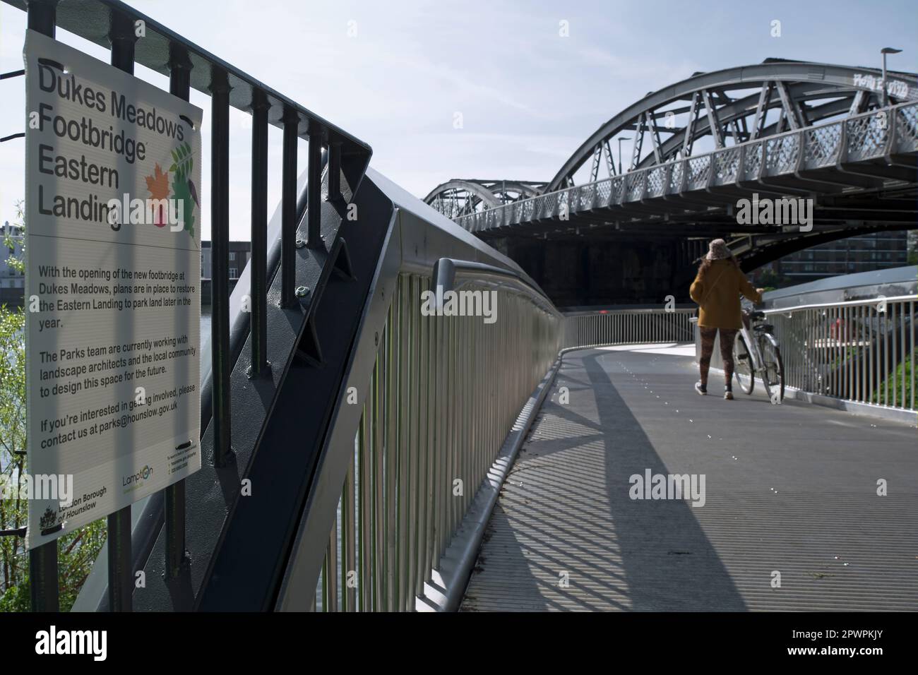 a woman pushes a bicycle on dukes meadows footbridge, below barnes bridge on the river thames, chiswick, london, england, with council notice beside Stock Photo