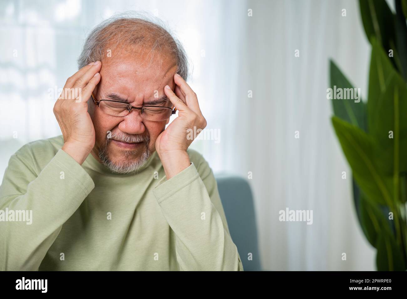 Headache. Sad Asian senior man sitting on sofa feeling hurt and lonely, elderly holds head with hand suffering from migraine headache, Old age health Stock Photo
