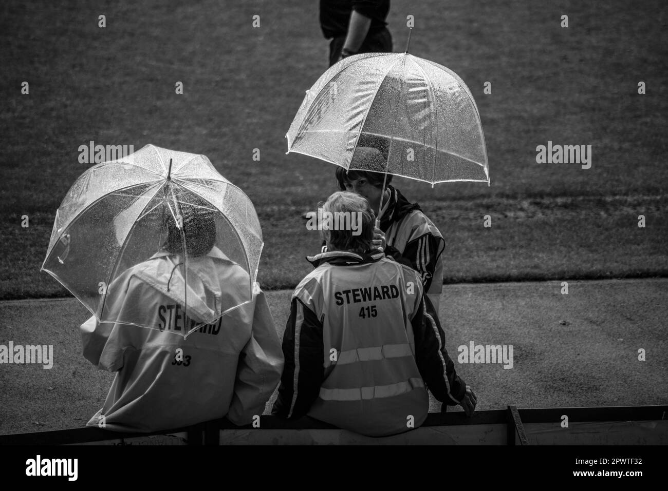 Black and white image of 3 stewards standing at the sidelines of a football match having a discussion using clear umbrellas at Upton Park, London Stock Photo