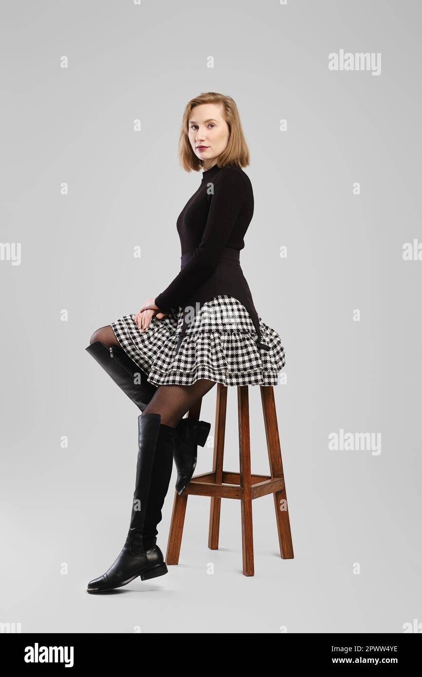 Young woman in turtleneck and suspender belt over little skirt sitting on tall chair in studio Stock Photo