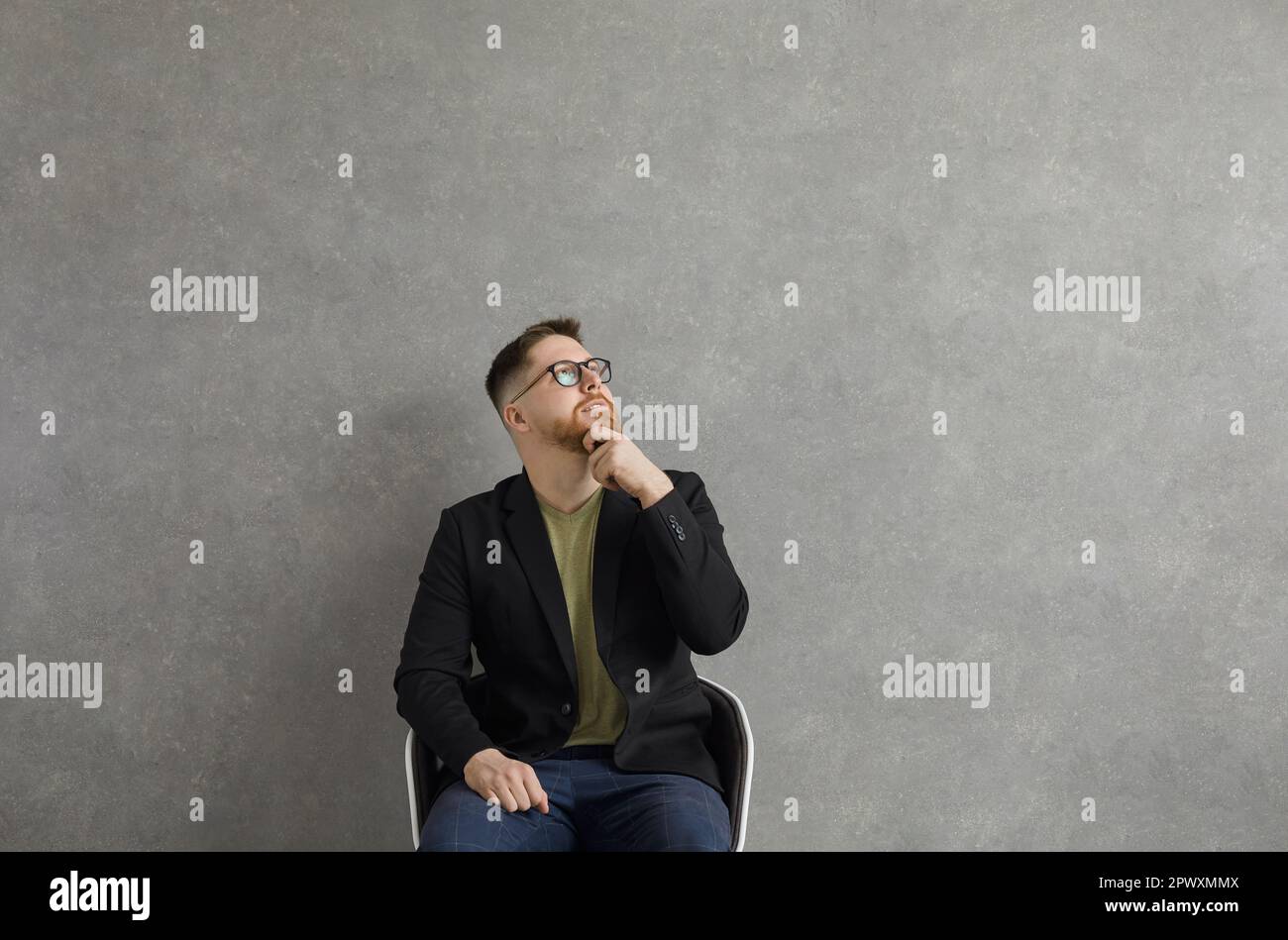 Serious young man in glasses sitting in chair, looking up and thinking about something Stock Photo