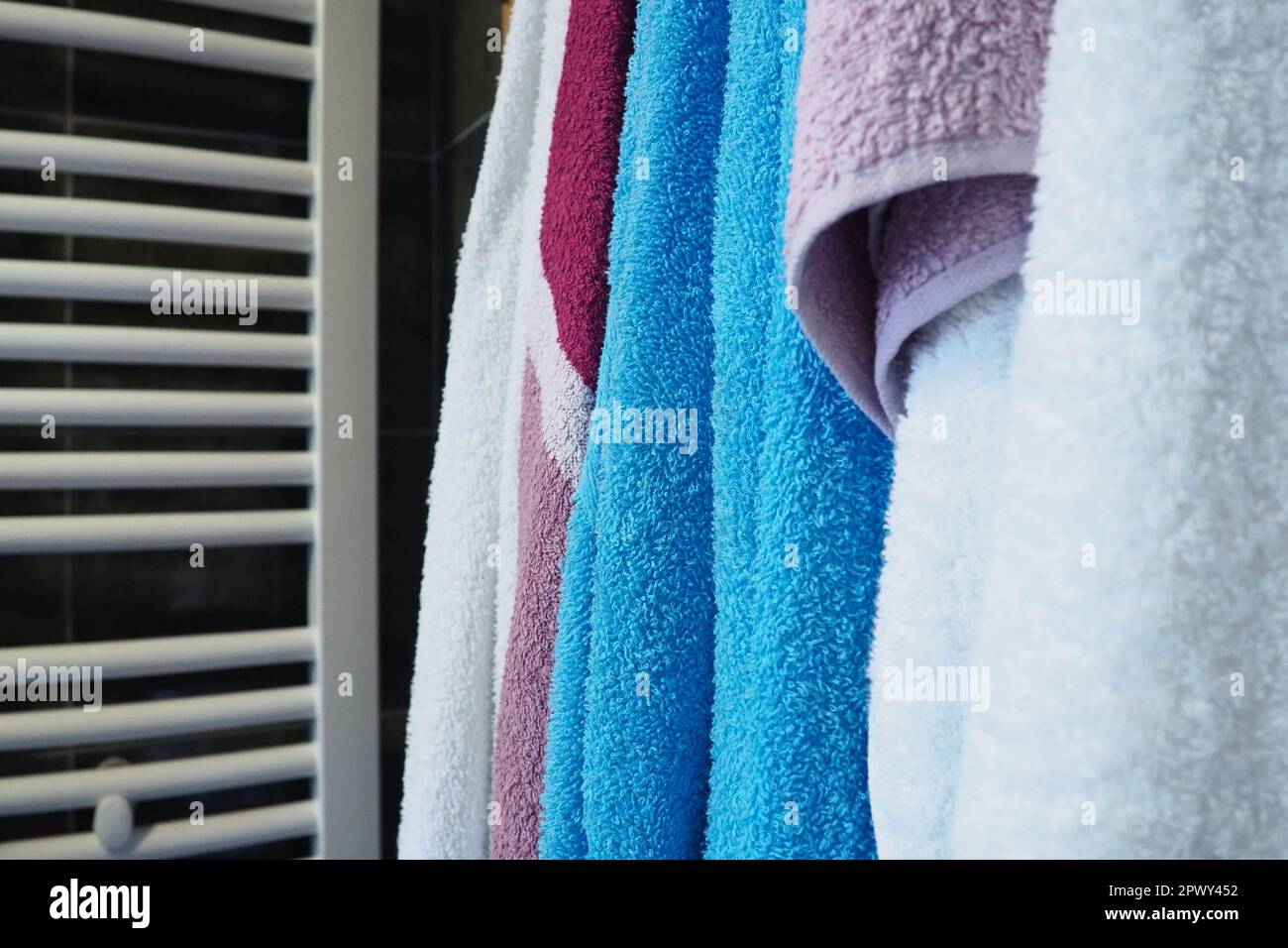 Towels hang next to a heated towel rail, wall radiator or radiator. White, blue, pink, red towels. Organization of household items in the bathroom. Ho Stock Photo