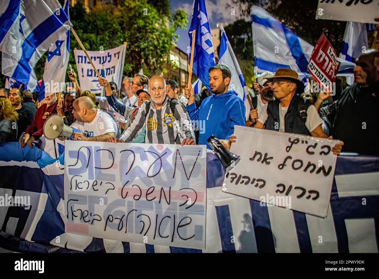 Protestors wave Israeli flags and hold placards against the Judicial system that read in Hebrew 'Bagatz (the Supreme Court) is killing the Jewish Majority' (R), and 'the Israeli Justice system is a holocaust to the people of Israel' as they demonstrate against former chief Justice Aharon Barak in Tel Aviv. Hundreds of pro and anti-judicial reform protesters gathered outside the home of former High Court Justice president Aharon Barak on Sunday, after anti-reform groups expressed concern for his safety earlier in the day. Stock Photo