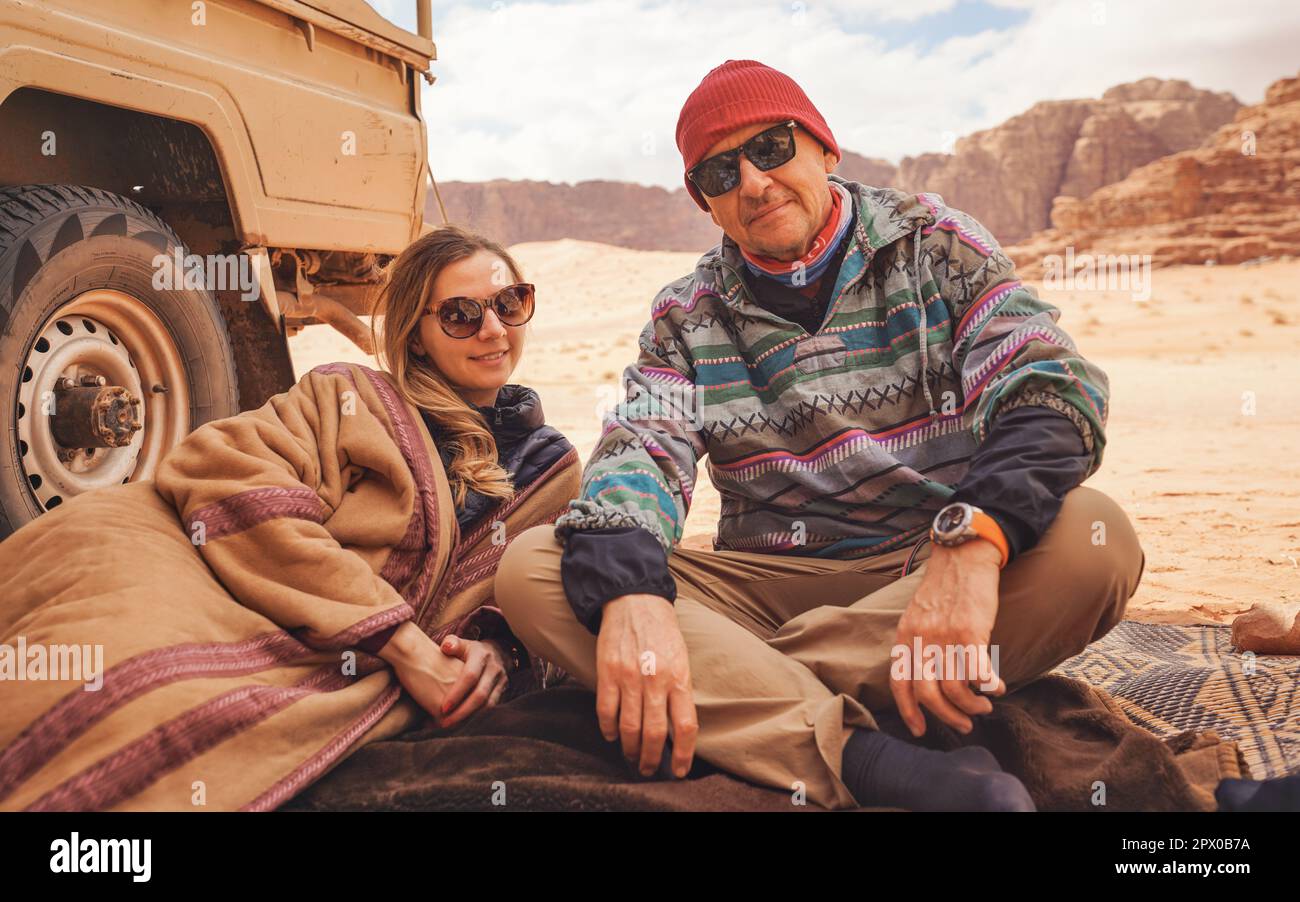 Man and younger woman wearing traditional Bedouin warm coat - bisht -  sitting or laying on ground blanket near off road vehicle back, blurred Wadi Ru Stock Photo