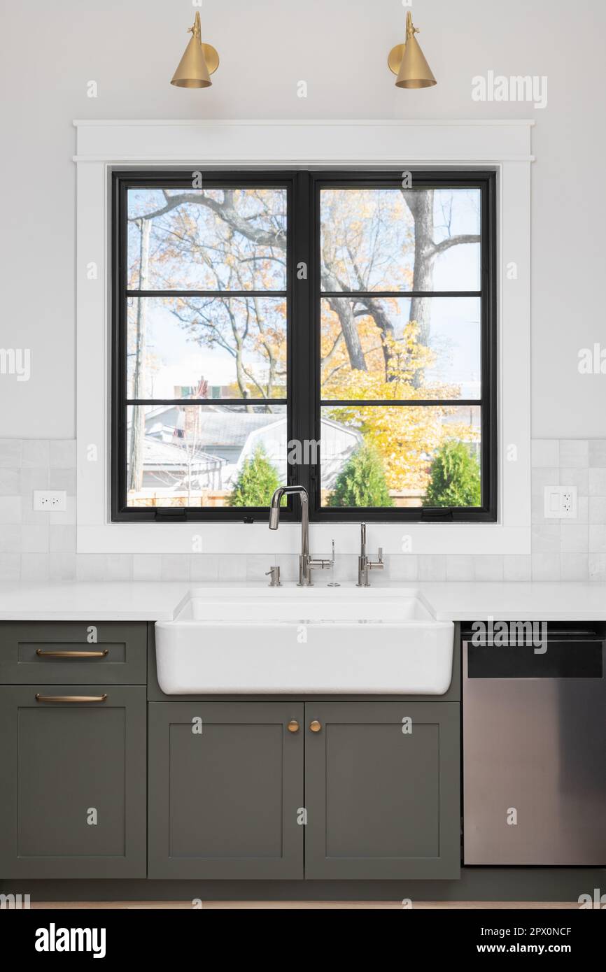 A sink detail shot with an apron sink, green cabinets, black window frame, a tiled backsplash, and gold sconces above the window. Stock Photo