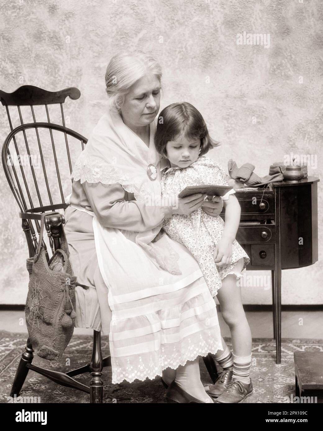 1930s SENIOR GRANDMOTHER SITTING IN WINDSOR CHAIR BY SEWING TABLE READING FROM SMALL BOOK TO HER LITTLE GRANDDAUGHTER - m2349 HAR001 HARS JUVENILE COMMUNICATION GENERATIONS EMBRACE STRONG GRANDPARENTS FAMILIES JOY LIFESTYLE FEMALES GRANDPARENT STUDIO SHOT HOME LIFE COPY SPACE FRIENDSHIP HALF-LENGTH HUG LADIES PERSONS INSPIRATION CARING EMBRACING SENIOR ADULT B&W SENIOR WOMAN QUALITY TIME YOUNG AND OLD GENERATION GRANDMOTHERS CONNECTION GRANDDAUGHTER STYLISH PERSONAL ATTACHMENT WINDSOR AFFECTION AGE DIFFERENCE ELDERLY WOMAN EMOTION GROWTH JUVENILES TOGETHERNESS BLACK AND WHITE Stock Photo