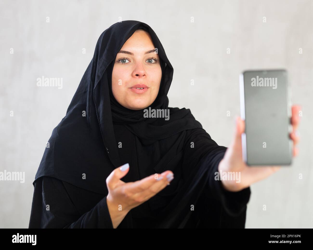 Cheerful young muslim lady in black traditional hijab showing new phone ...