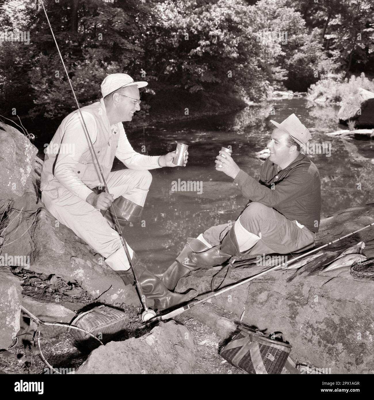 1960s TWO MEN FISHERMEN BY SIDE OF STREAM TOASTING EACH OTHER ON DAY’S ...