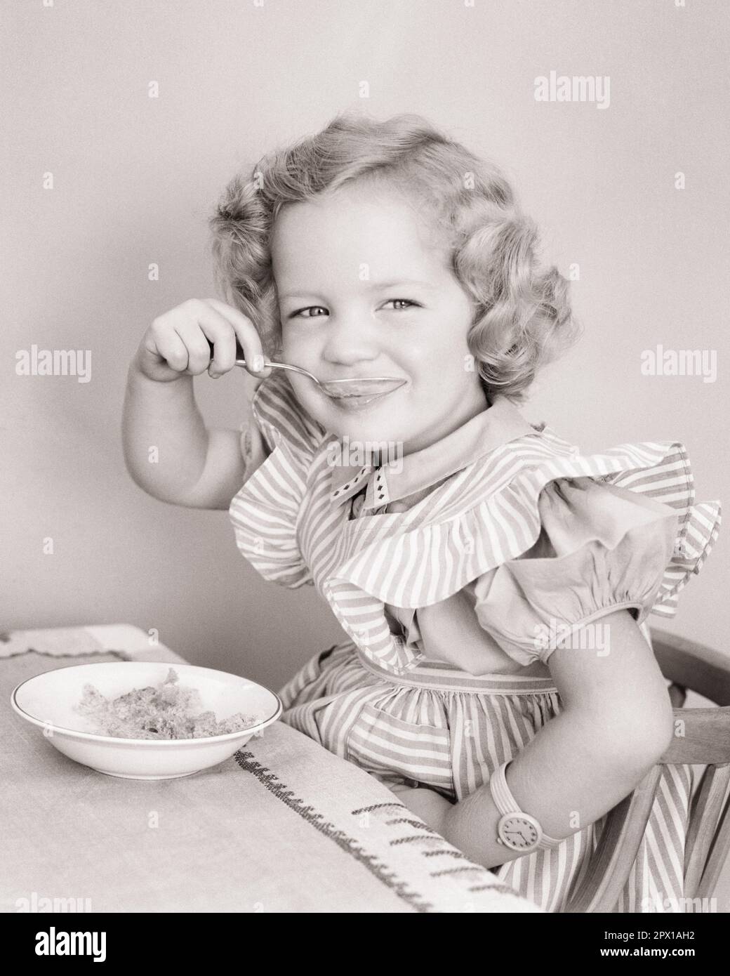 1940s CUTE SMILING LITTLE GIRL IN FANCY DRESS LOOKING AT CAMERA EATING BREAKFAST CEREAL - f2242 HAR001 HARS HALF-LENGTH FANCY STRIPES CONFIDENCE B&W EYE CONTACT HAPPINESS WELLNESS NUTRITION CONSUME CONSUMING NOURISHMENT STYLISH PINAFORE PLEASANT AGREEABLE CHARMING COOPERATION GROWTH JUVENILES LOVABLE PLEASING ADORABLE APPEALING BLACK AND WHITE CAUCASIAN ETHNICITY HAR001 OLD FASHIONED Stock Photo