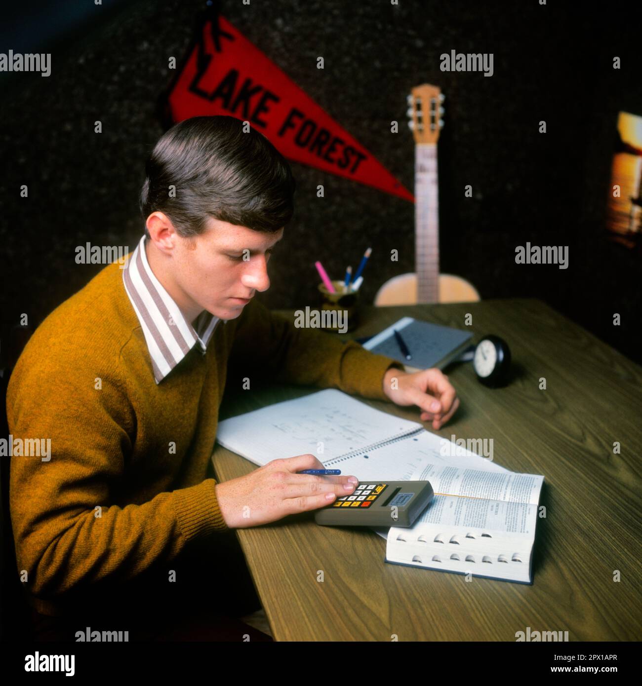 1970s MALE COLLEGE STUDENT STUDYING AT DESK USING REFERENCE BOOK CALCULATOR TAKING NOTES DORM ROOM GUITAR PENNANT IN BACKGROUND - ks14368 BAN001 HARS INFORMATION LIFESTYLE HOME LIFE COPY SPACE HALF-LENGTH PERSONS THOUGHTFUL MALES TEENAGE BOY DORM GOALS SCHOOLS REFERENCE UNIVERSITIES HIGH SCHOOL PENNANT USING HIGH SCHOOLS HIGHER EDUCATION TEENAGED COLLEGES SOLEMN FOCUSED GROWTH INTENSE JUVENILES YOUNG ADULT MAN CAREFUL CAUCASIAN ETHNICITY EARNEST INTENT OLD FASHIONED Stock Photo