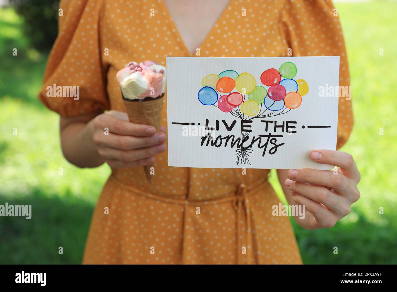 Woman holding card with phrase Live The Moments and ice cream outdoors, closeup Stock Photo