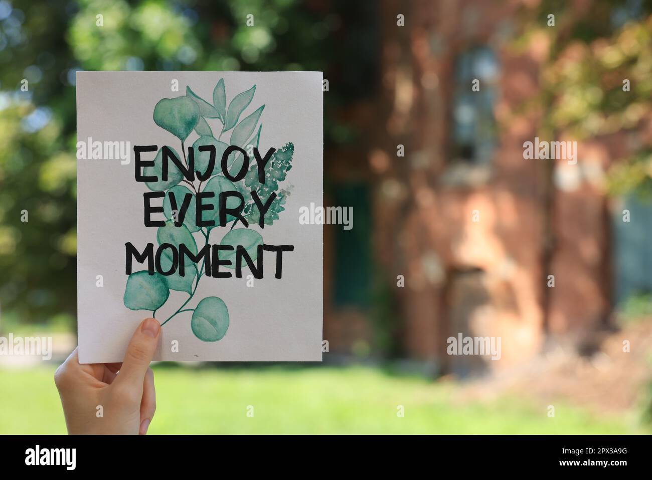 Woman holding card with phrase Enjoy Every Moment outdoors, closeup. Space for text Stock Photo