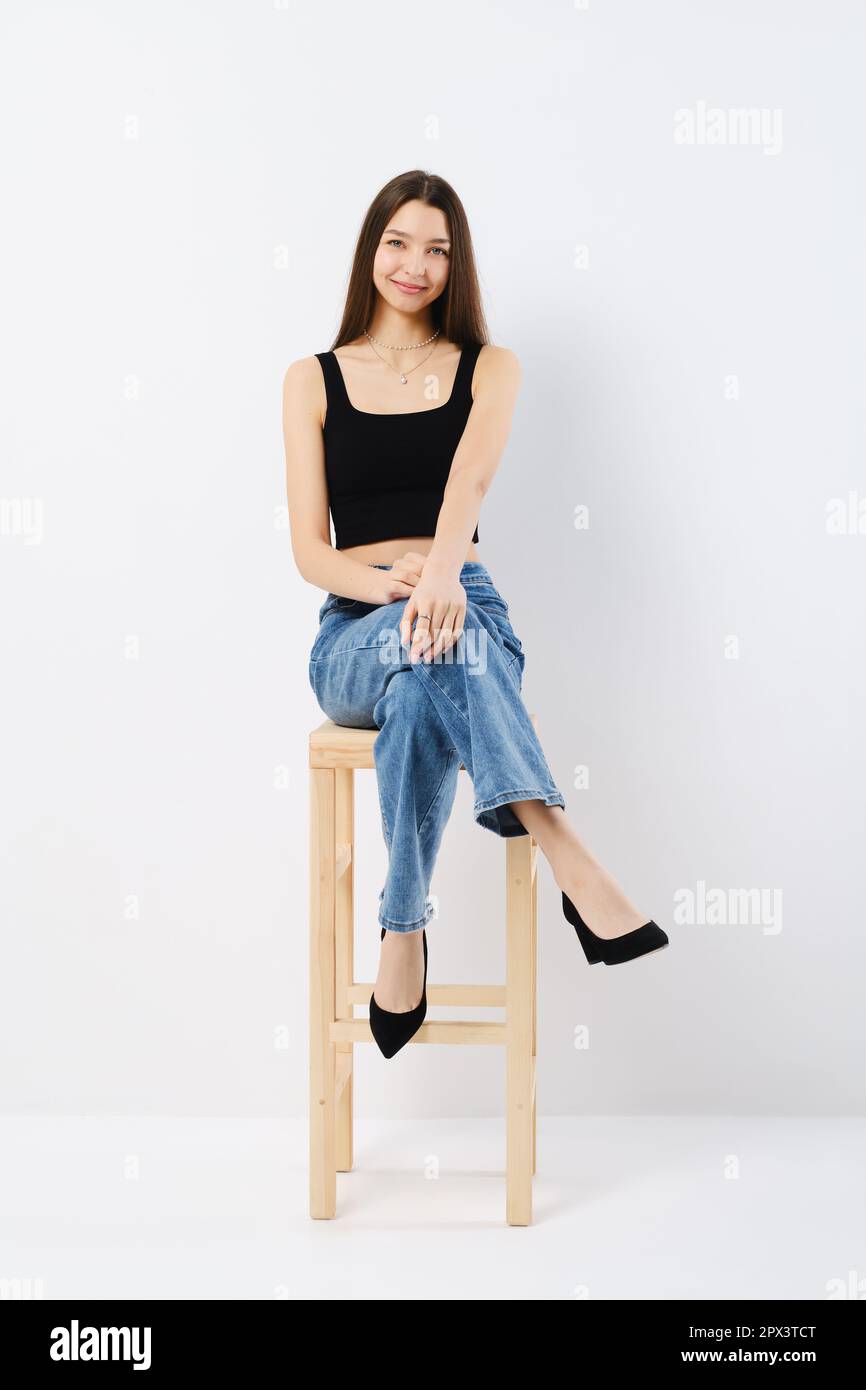 Smiling young dark-haired girl dressed in a black top, shorts, tights and  cap is sitting on the floor on the white background in the studio Stock  Photo