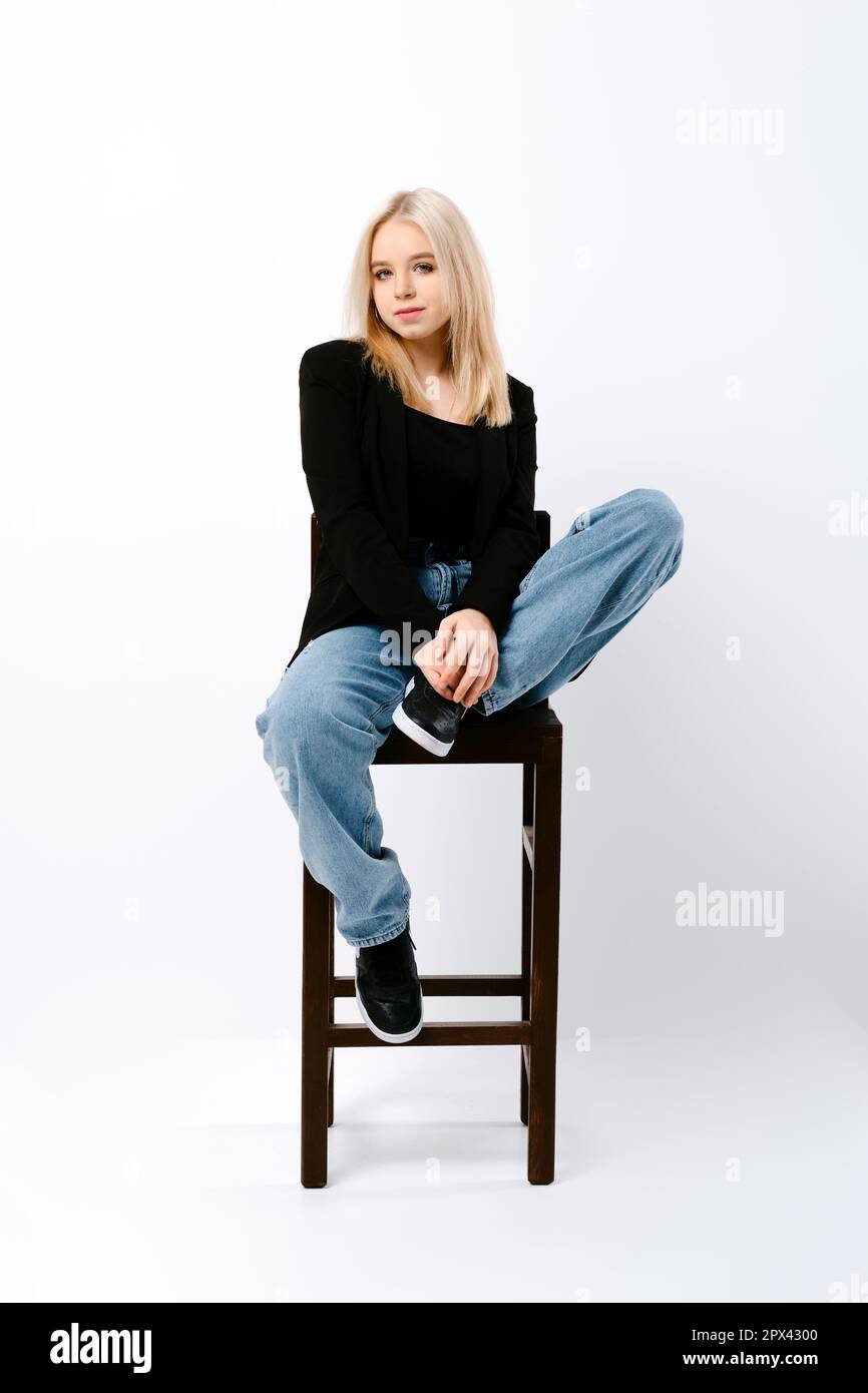 Teen girl in tank top, jacket and jeans posing on chair in white studio