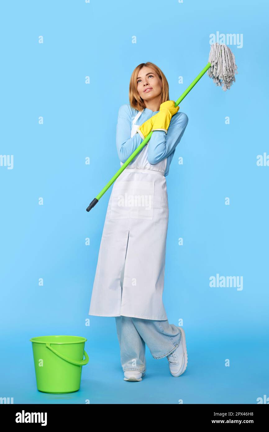 professional cleaner blonde woman in rubber gloves and cleaner apron washing floor with mop on blue background. Full length Stock Photo