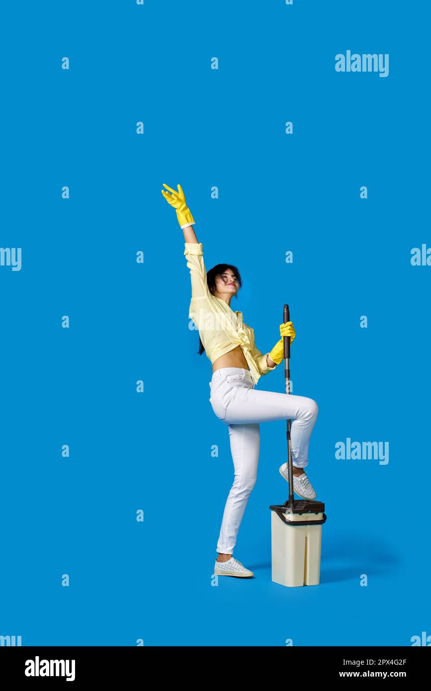 funny happy housewife girl in yellow rubber gloves washing floor with mop and dancing on blue background. Full length Stock Photo