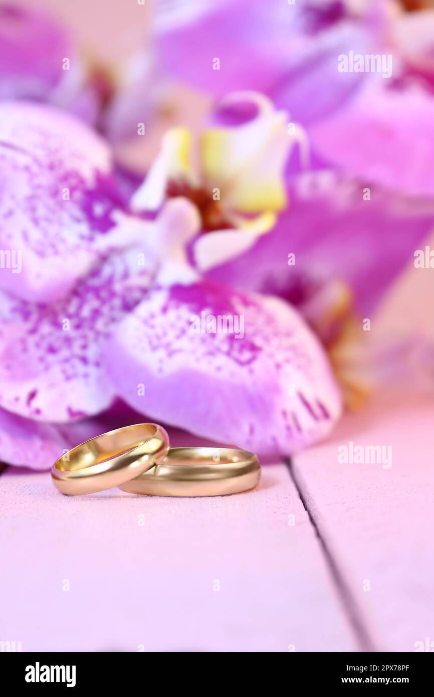Golden wedding rings with purple orchids on pink wooden table. Vertical. Wedding ceremony, honeymoon, marriage proposal, Valentine's day in hot countr Stock Photo