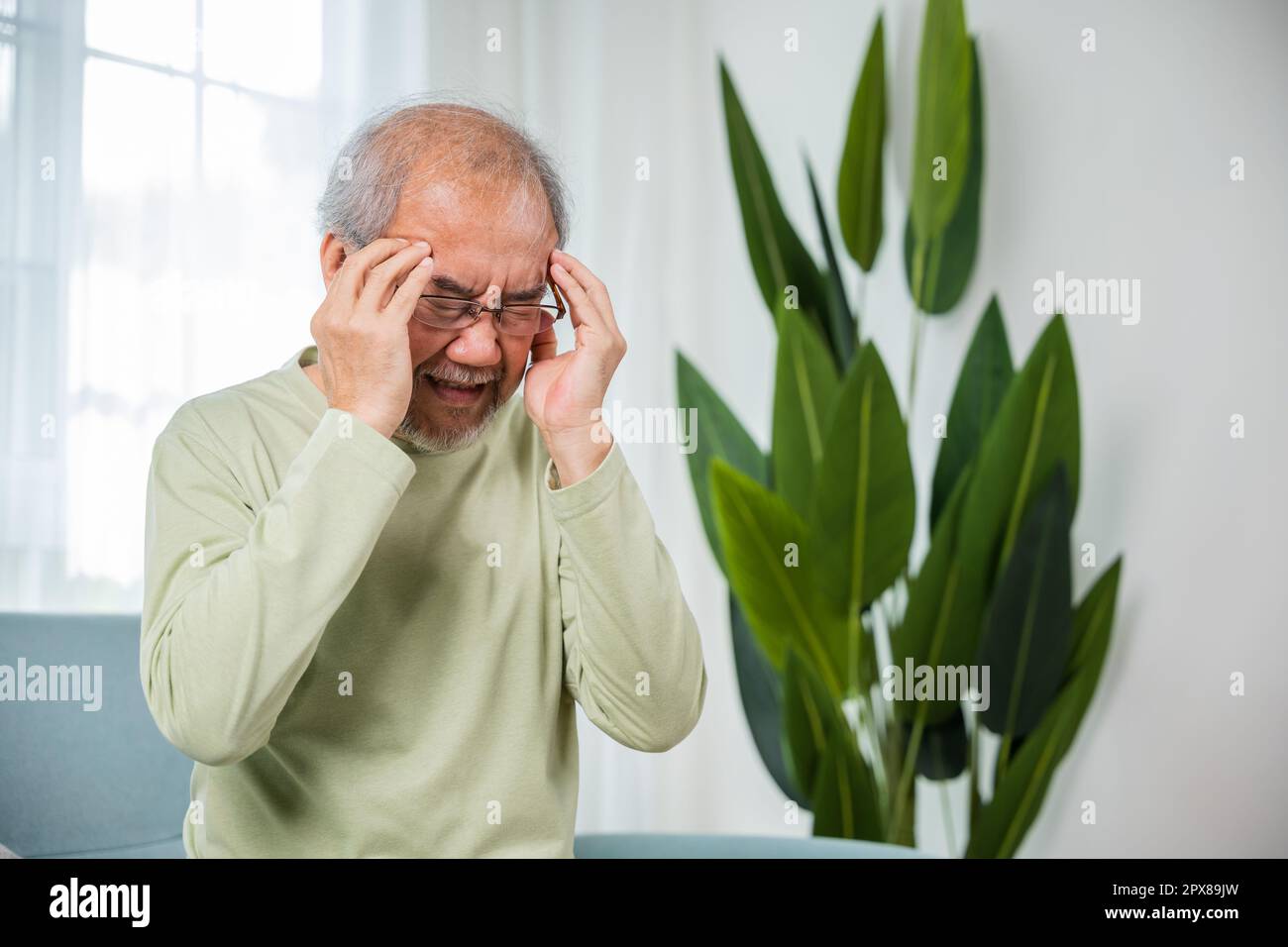 Headache. Sad Asian senior man sitting on sofa feeling hurt and lonely, elderly holds head with hand suffering from migraine headache, Old age health Stock Photo