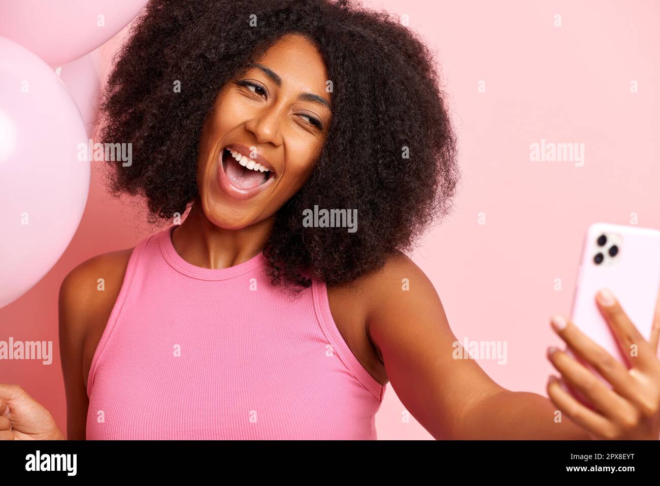 Charming dark skinned woman holds a smartphone and makes a selfie, smiles happy and widely, isolated next to pink wall. High quality photo Stock Photo