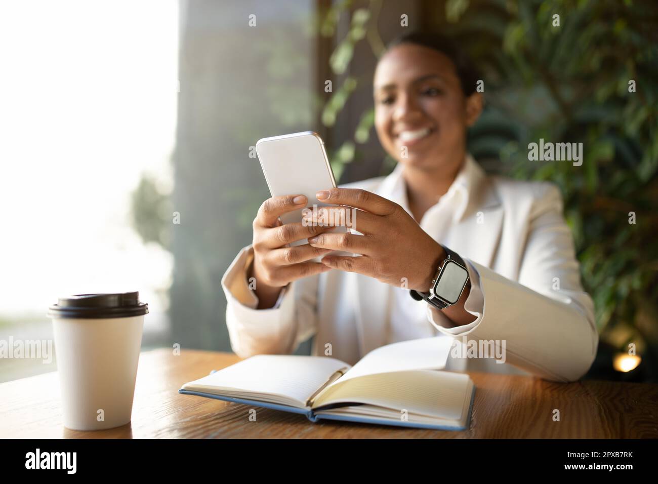 Positive young black lady in white suit at table chatting on phone, with cup of coffee and notebook in cafe Stock Photo
