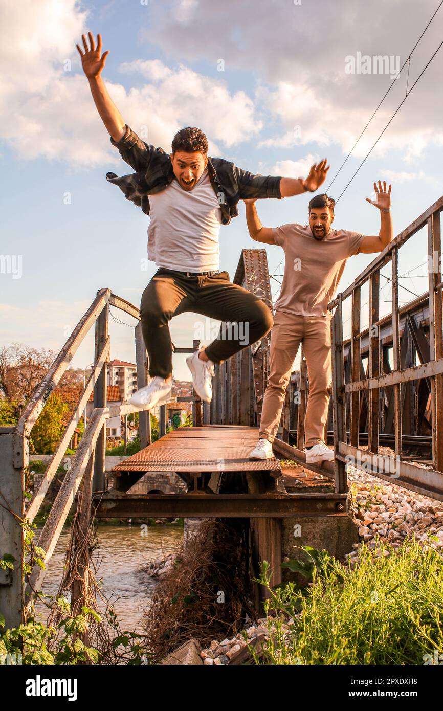 Caucasian teenager jumping from train tracks on a rusty train bridge with a blurry city in the background and his friend watching him Stock Photo