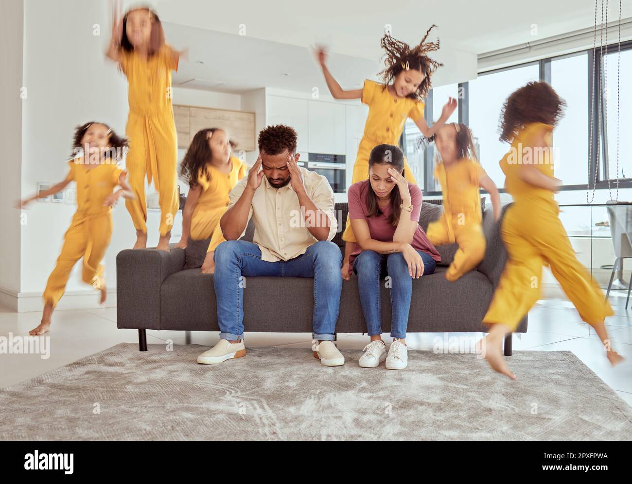 Stressed mixed race parents and adhd daughter in living room at home. Hyperactive hispanic girl running and jumping around lounge. Mother and father Stock Photo