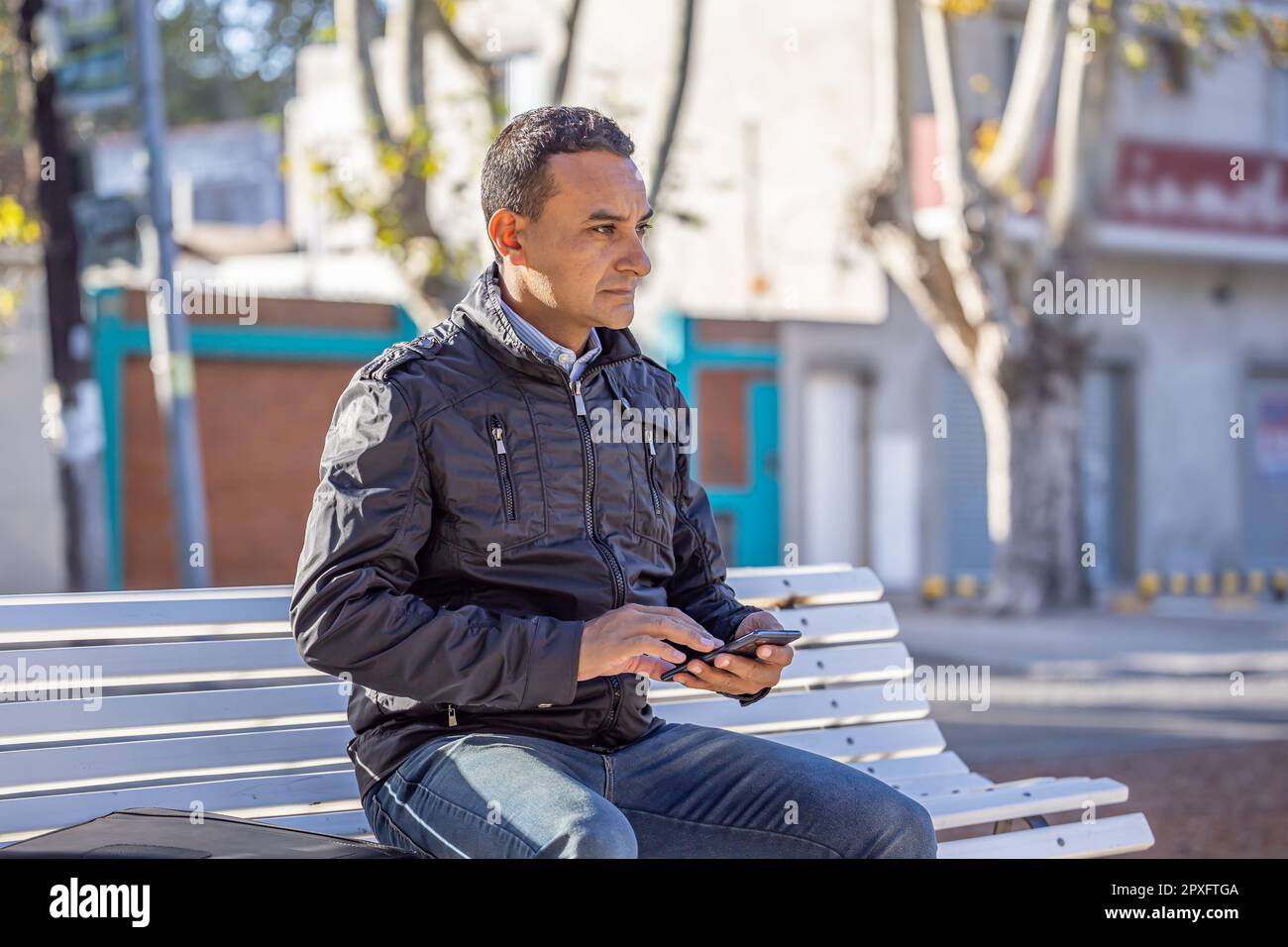 Young latin man sitting on a square bench using his mobile phone. Stock Photo