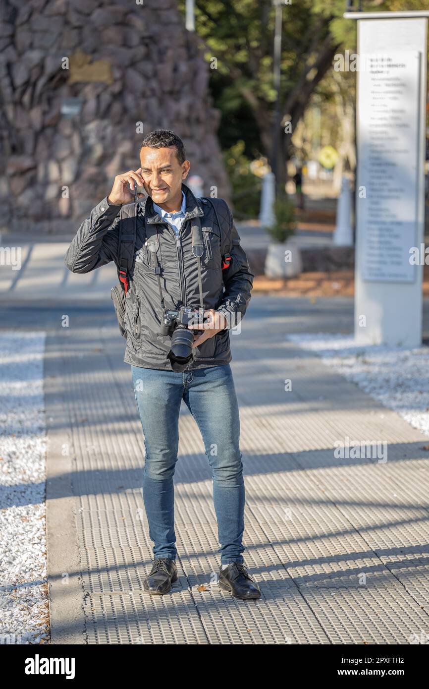 Young latin man talking on mobile phone with a photo camera around his neck. Stock Photo