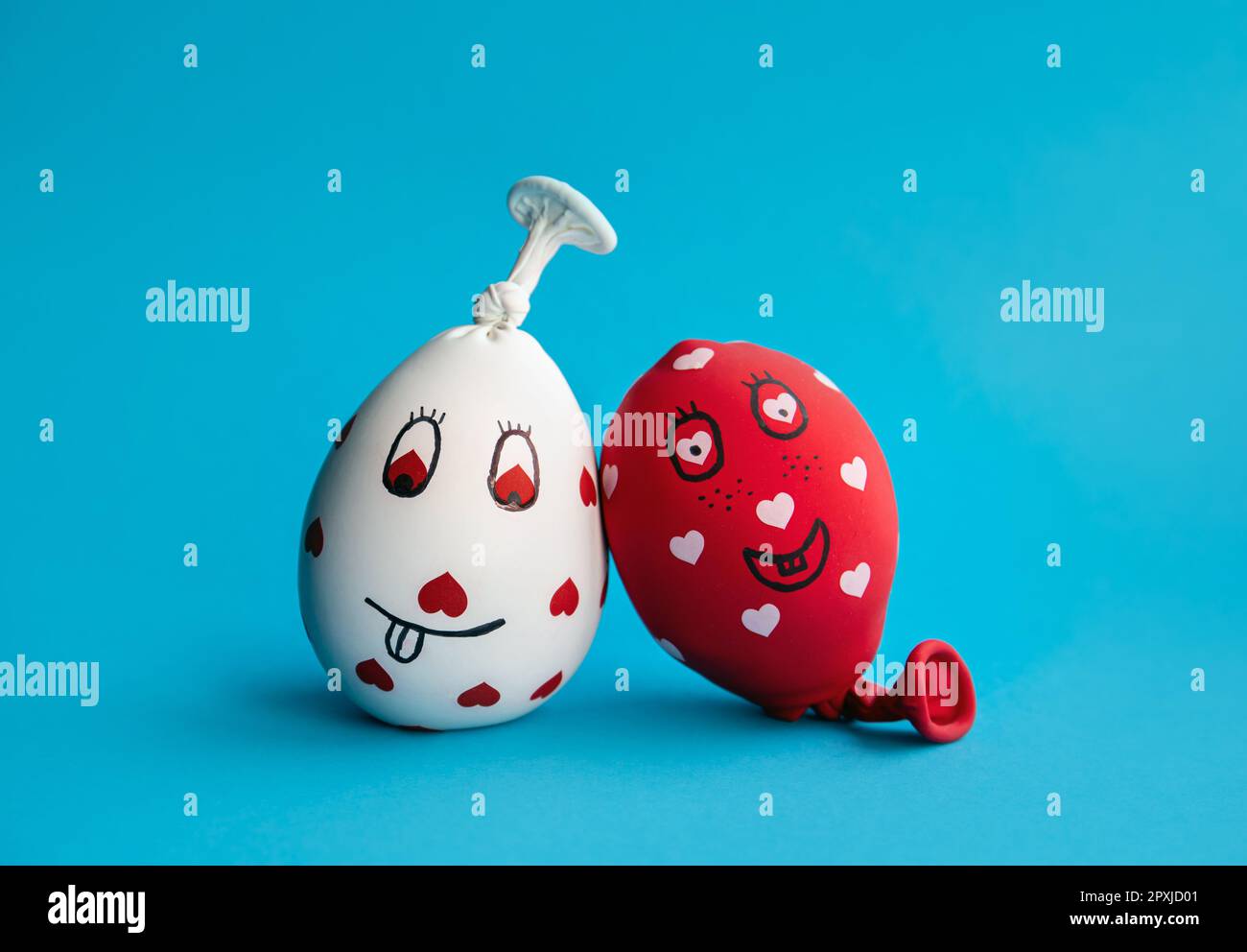 Two funny homemade sensory stress balls, made of balloons and filled with flour. Red and white balls with hearts and funny drawn faces on blue minimal Stock Photo