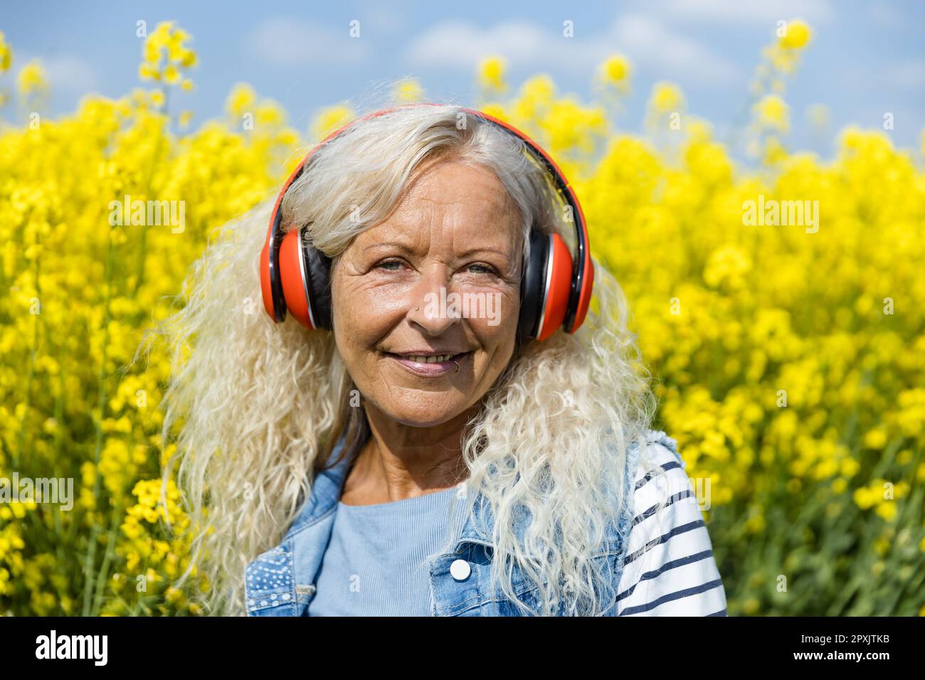 Elderly Woman listen to music with  headphones in blooming canola field Stock Photo