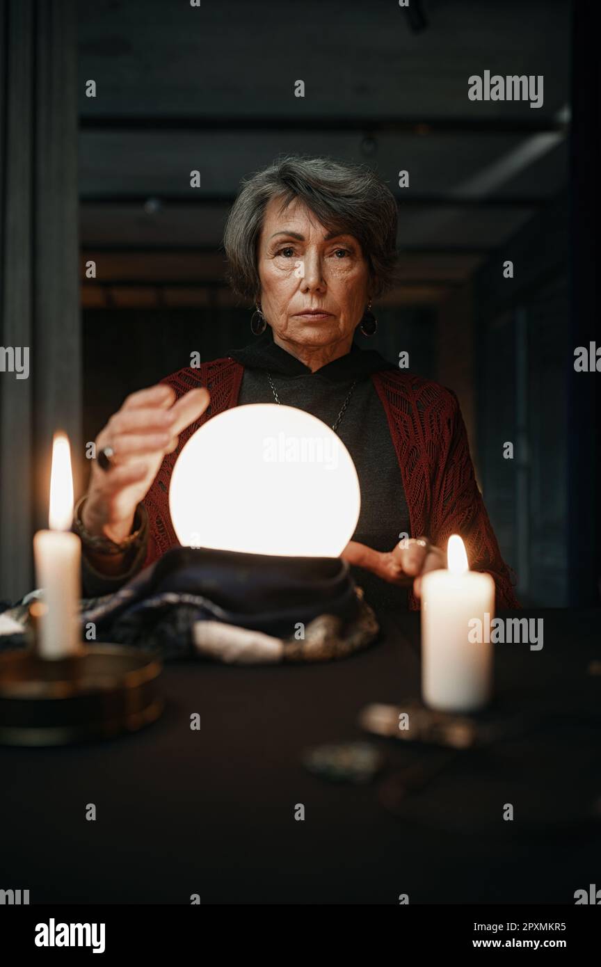 Confident witch fortune teller doing predictions with illuminated crystal ball reading future during esoteric ritual Stock Photo