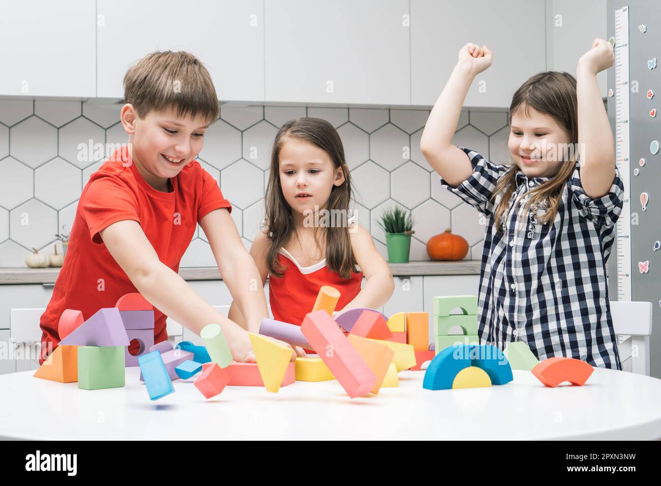 Portrait of happy preteen children friends sitting at table in kitchen, breaking tower house castle of colorful wooden construction blocks geometric f Stock Photo
