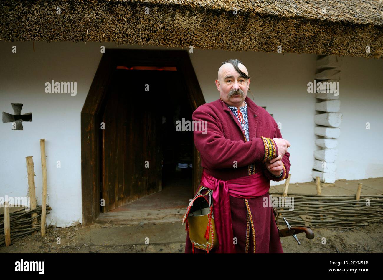 Man in Ukrainian native costume of cossack and saber standing in front of his rural house. Ukrainian folk traditions, reconstruction. January 4, 2018. Stock Photo