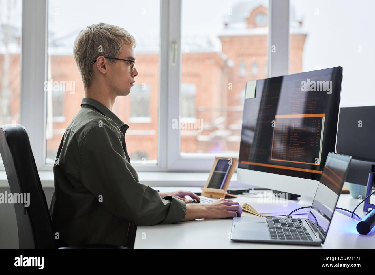Side view of young programmer sitting at his workplace and working with security codes for program using computer and smartphone Stock Photo