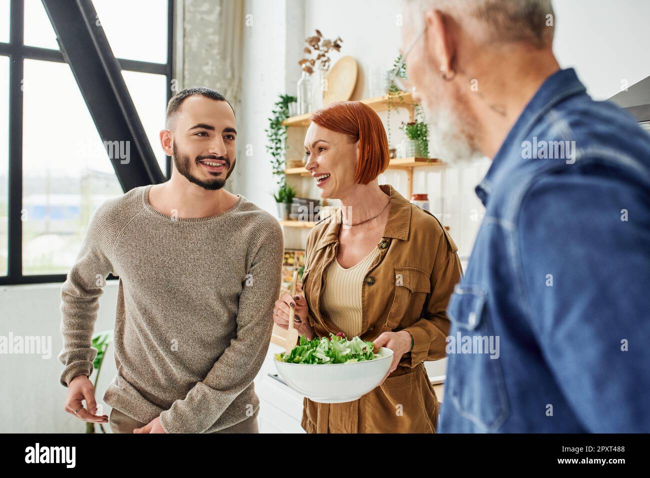 happy redhead woman holding bowl with fresh salad near son and blurred husband in kitchen,stock image Stock Photo