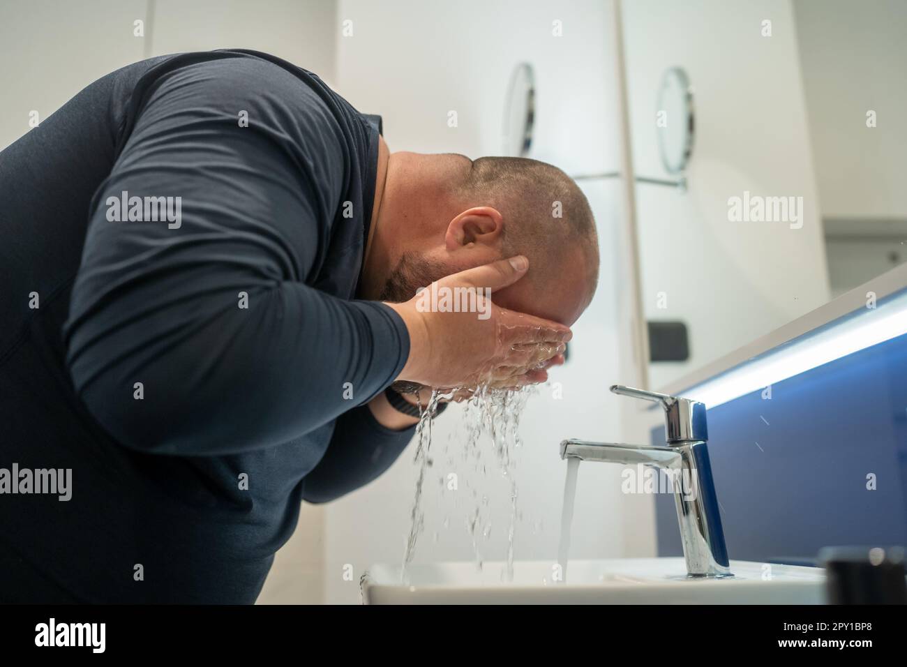 Overweight balded man washing face in bathroom at home under sink, side