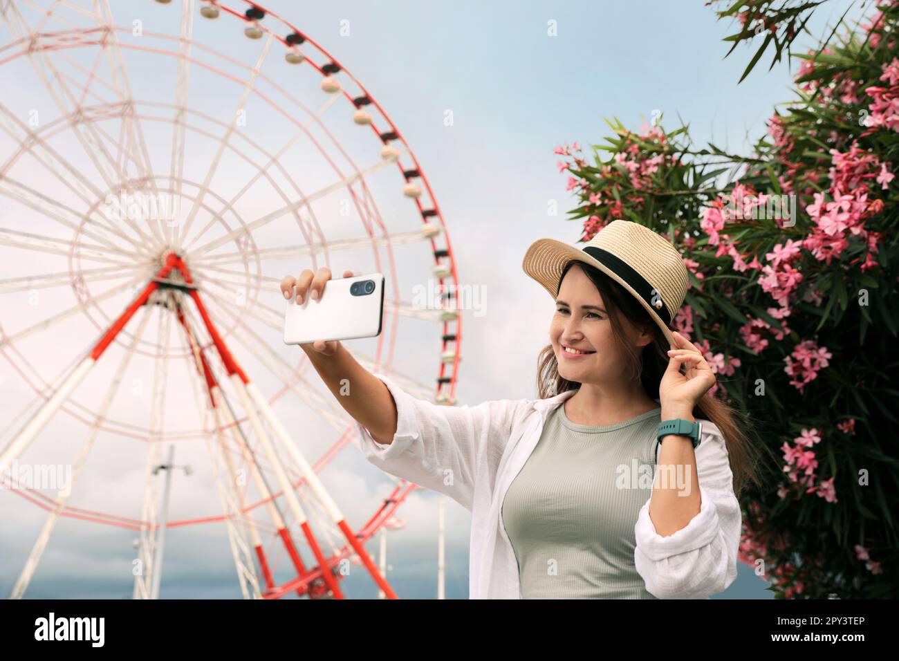Woman taking selfie near beautiful blooming oleander and large Ferris wheel outdoors Stock Photo