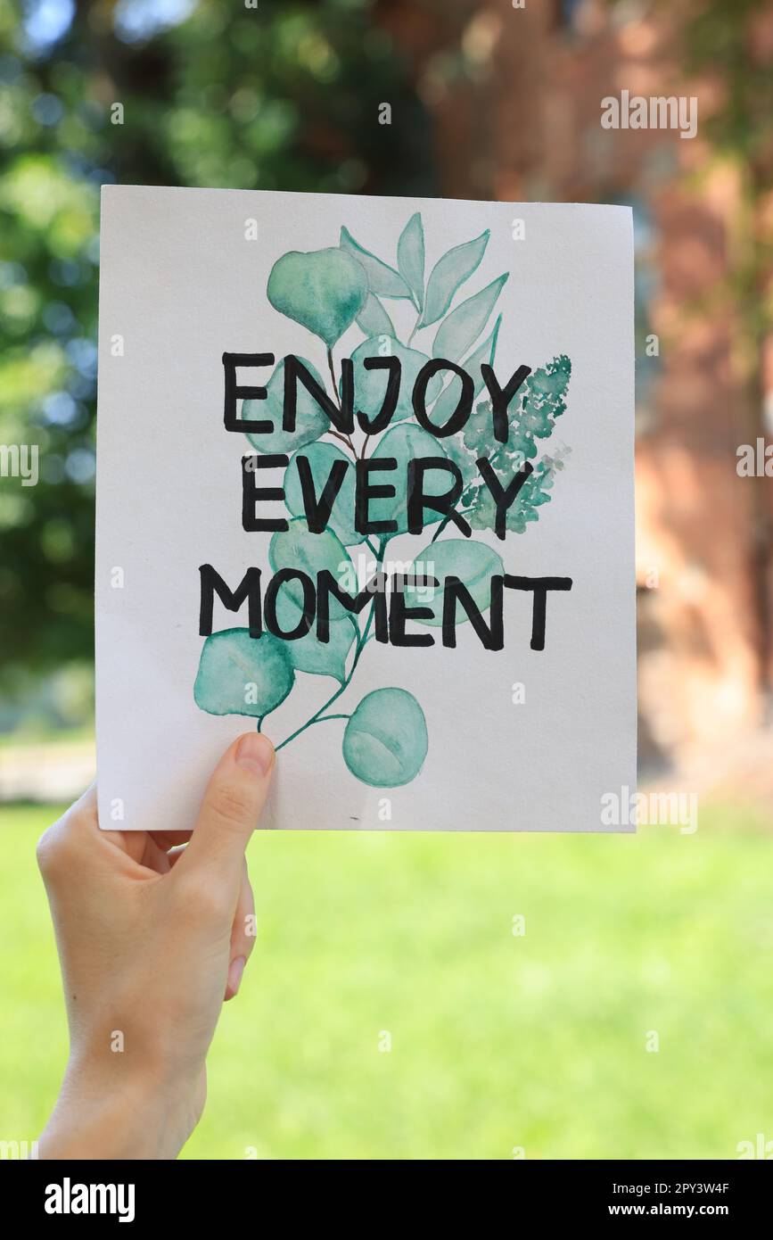 Woman holding card with phrase Enjoy Every Moment outdoors, closeup Stock Photo