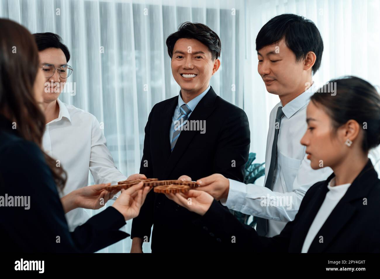 Closeup hand holding wooden gear by businesspeople wearing suit for harmony synergy in office workplace concept. Group of people hand making chain of Stock Photo