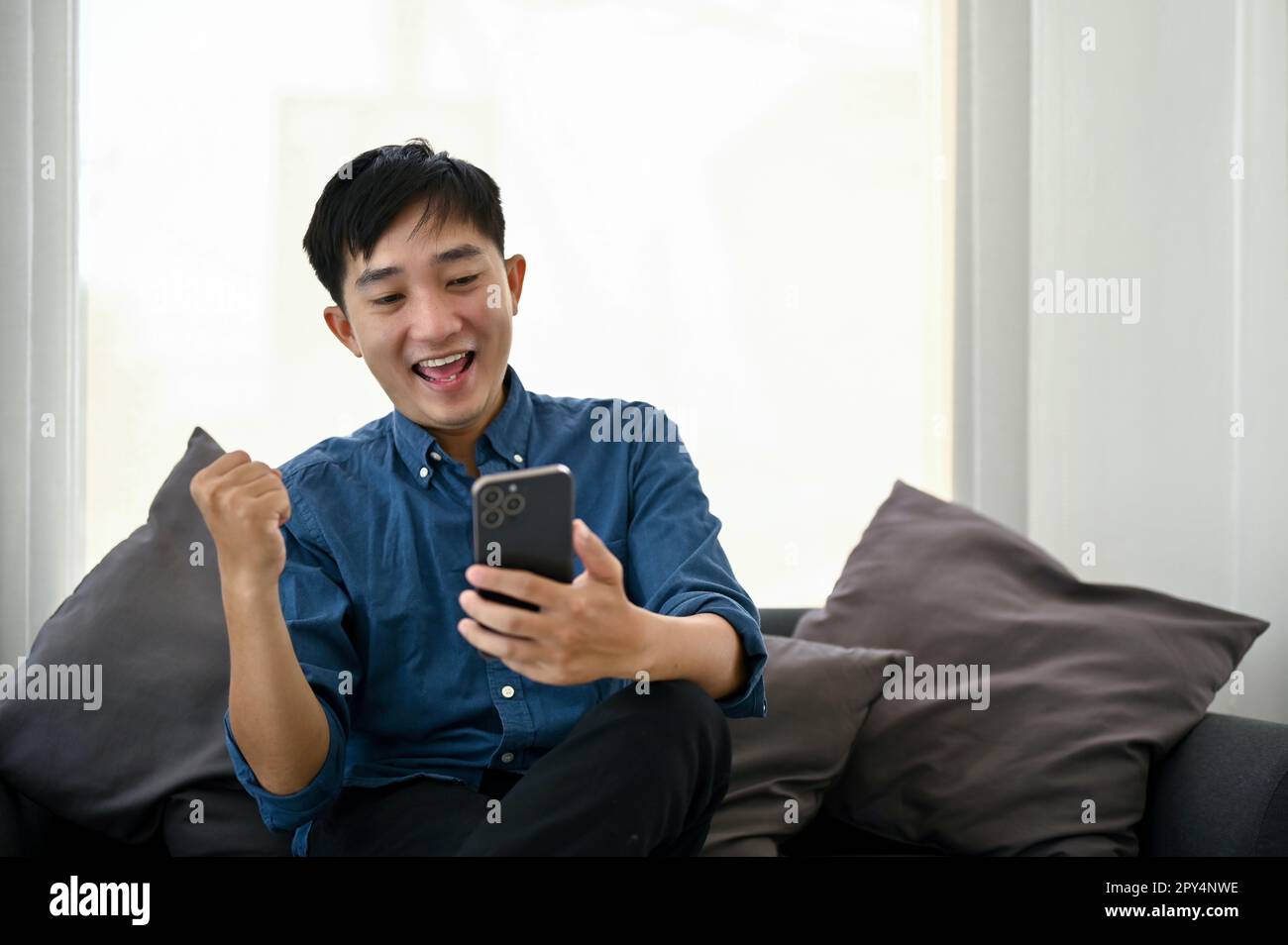 Cheerful millennial Asian man celebrating good news after get an unexpected sms or email via his smartphone, sitting on sofa in the living room. Stock Photo