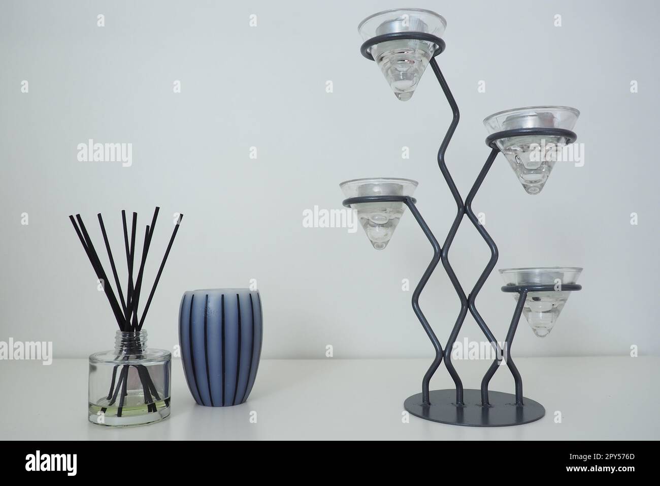 Tall metal chandelier with glass holders for small candles. White background. Incense sticks in a glass cubic vessel with essential oil. Interior decor. Scented gray striped candle or vase. Stock Photo