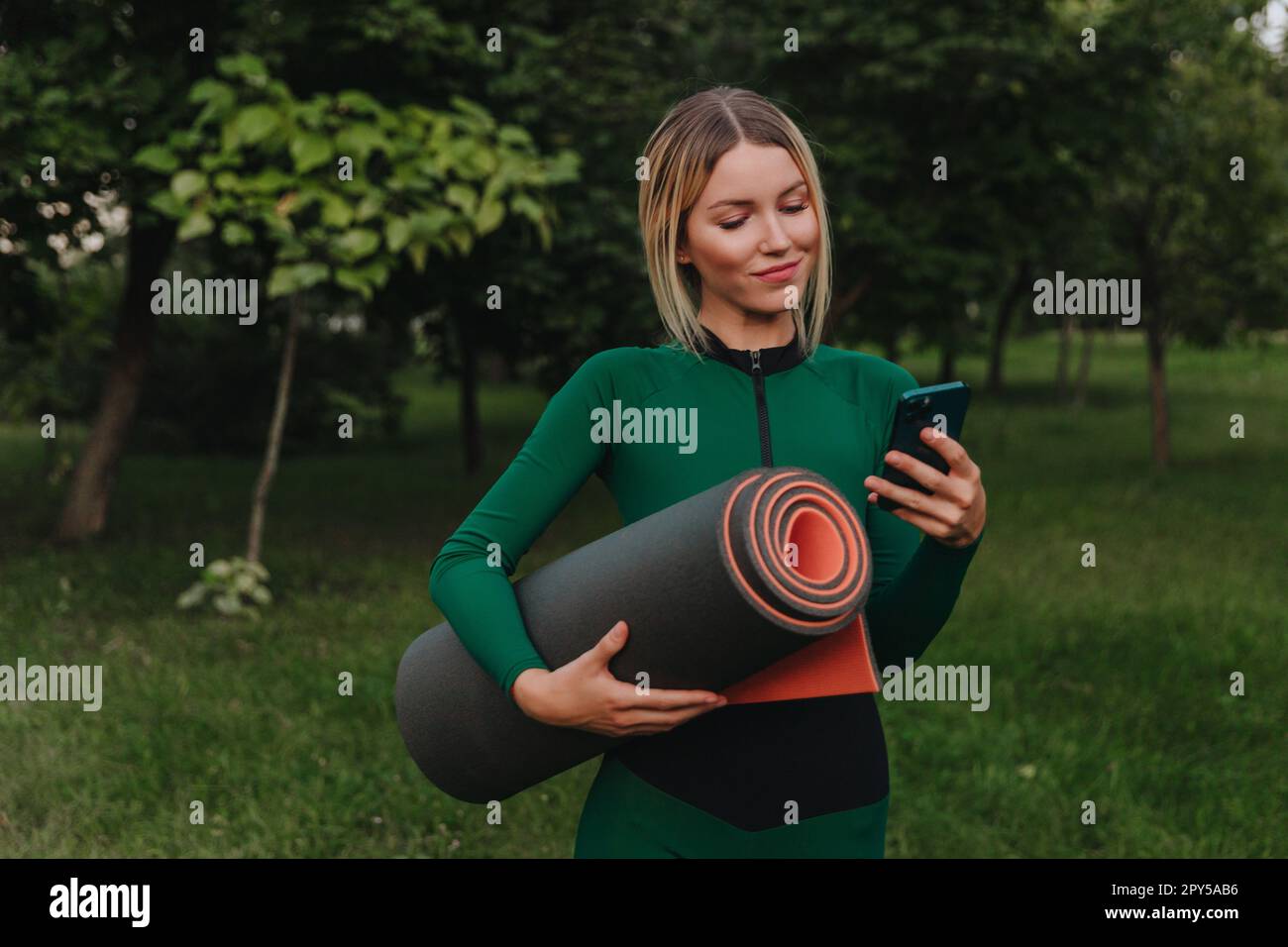 Yoga in the park with sunlight. A young woman in a lotus position sits on green grass holding a phone in her hand. The concept of digital transformation. Stock Photo