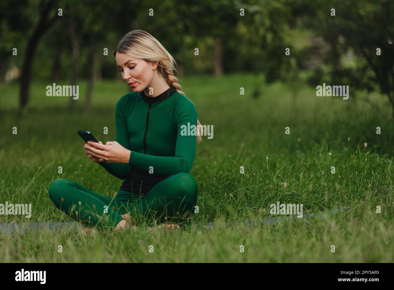Yoga in the park with sunlight. A young woman in a lotus position sits on green grass holding a phone in her hand. The concept of digital transformation. Stock Photo