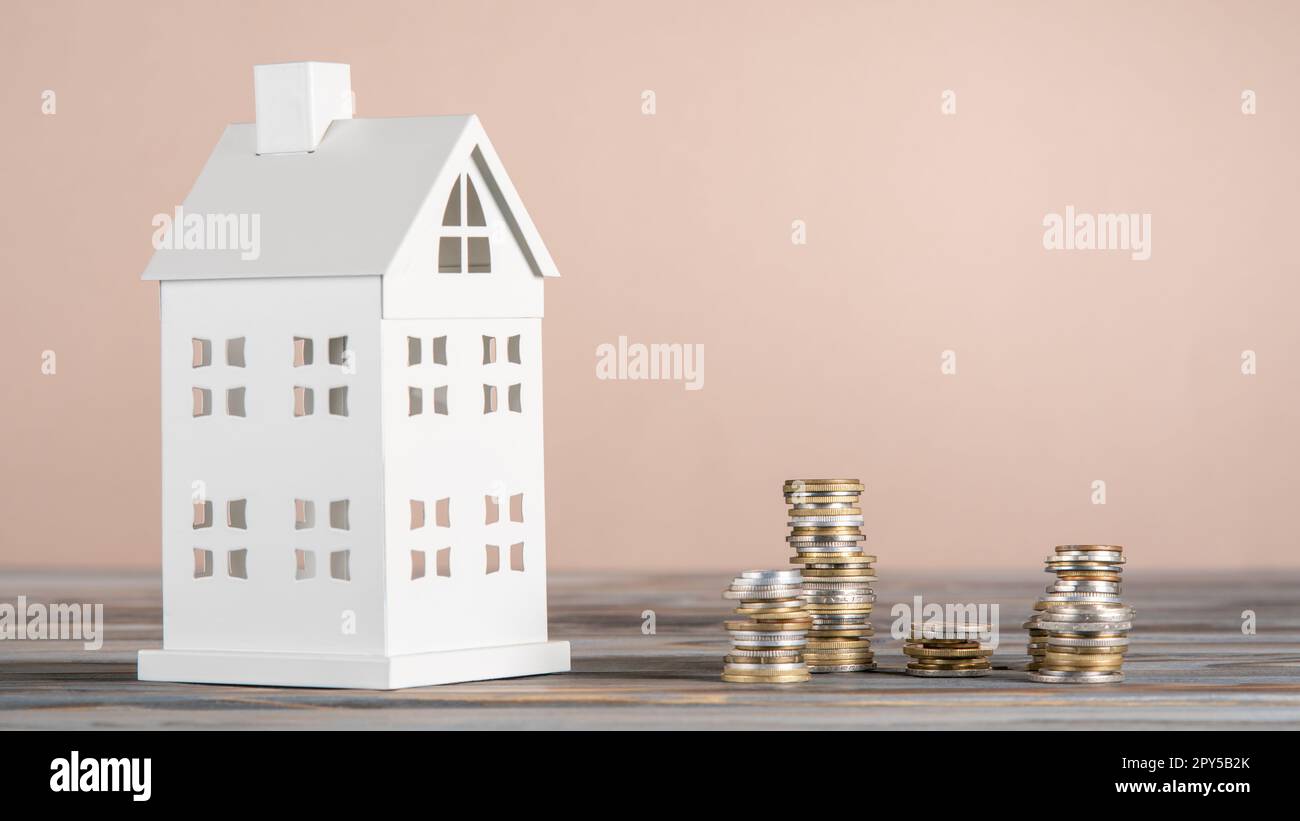 White toy model of house with white stacks of coins made of carton paper on grey table on pink background. Investment. Stock Photo