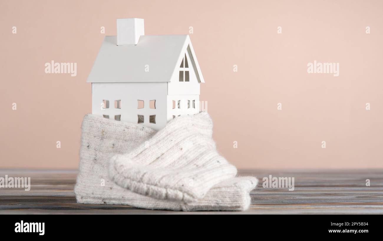 White toy model of house with white knitted scarf made of carton paper on grey table on pink background. Heating system. Stock Photo