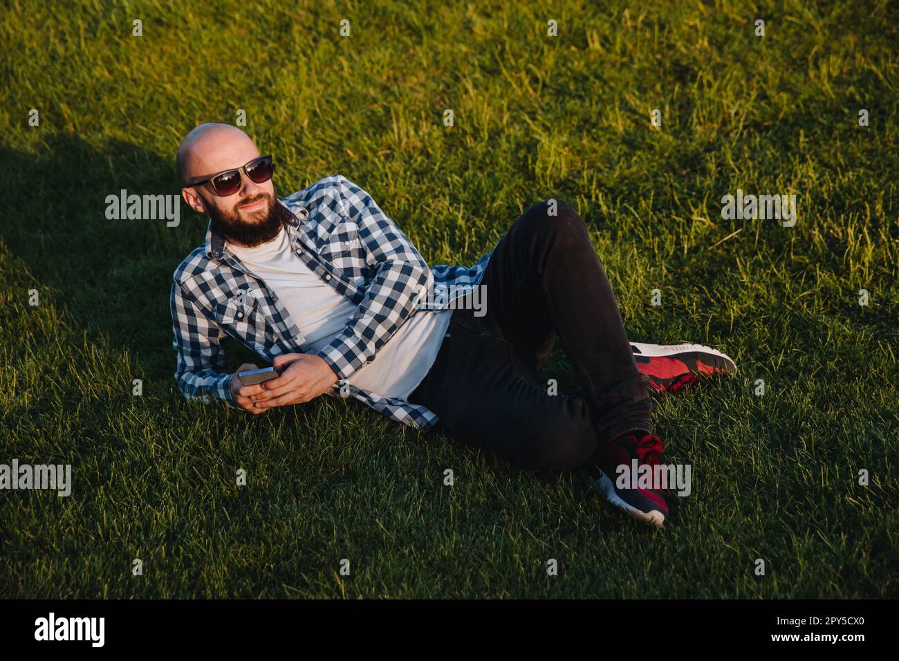 A young man sits on the green grass in the park and uses social networks using a mobile phone. Stock Photo