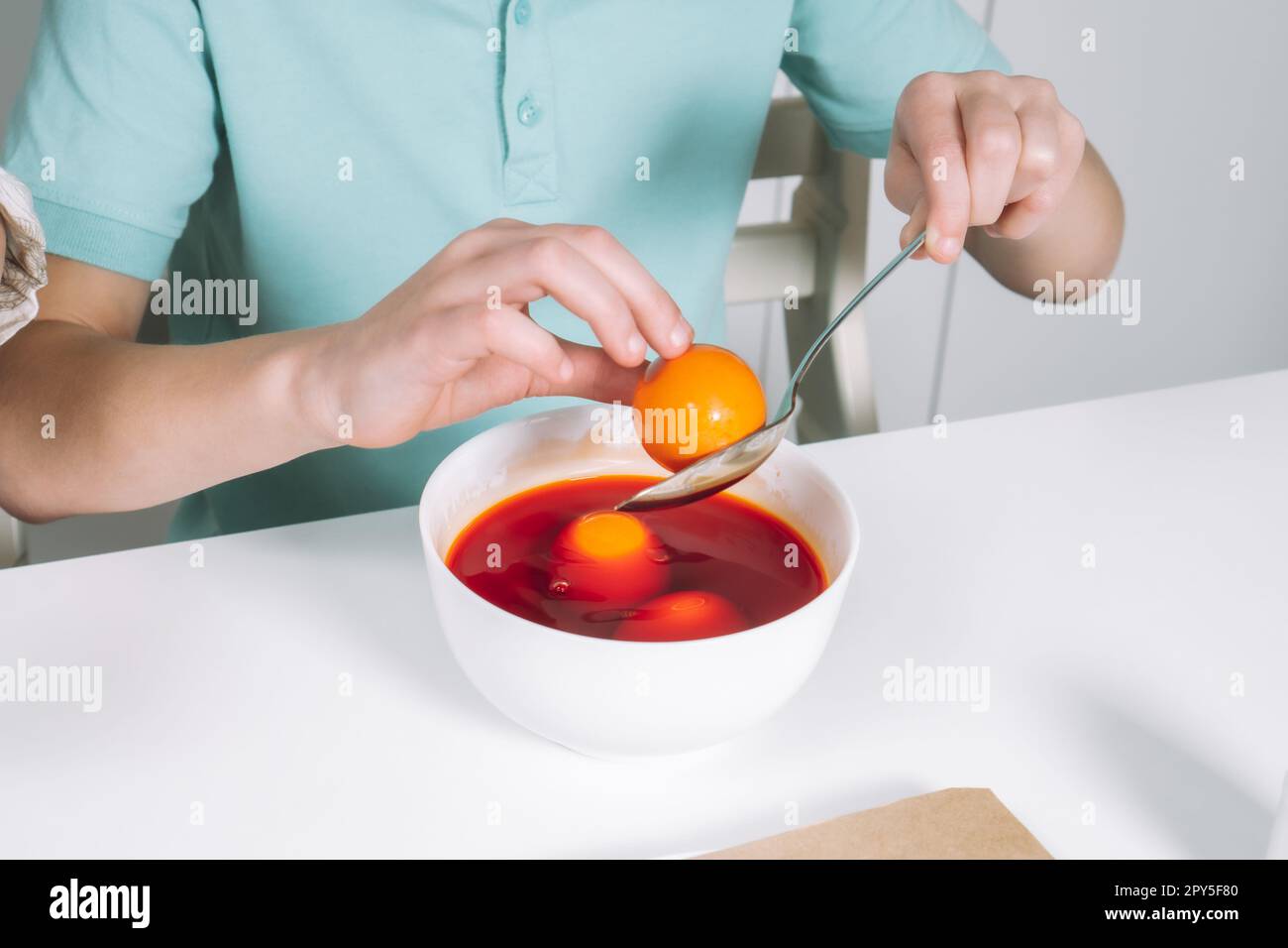 Cropped photo of boy hands, coloring orange Easter hen eggs in spoon, bowl, table. Soak up, absorption color. Close up Stock Photo